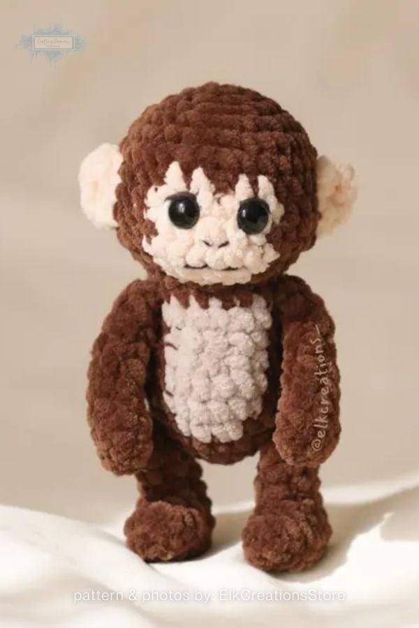An image of a crochet monkey plushie from a crochet roundup on craftinghappiness.com. The monkey is made from soft, chunky brown yarn with a cream-colored face, ears, and belly. It features large, dark safety eyes and a simple embroidered mouth, standing upright against a neutral background.