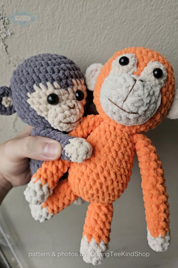 An image of two handmade crochet monkey plushies from a crochet roundup on craftinghappiness.com. Held in a person's hand, the pair includes a larger orange monkey and a smaller grey one, both made from textured yarn with cream-colored faces and paws. They feature round black safety eyes and simple embroidered smiles, posed against a neutral, textured wall background.