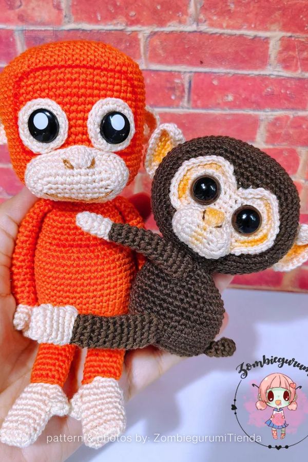 An image of two handmade crochet monkey plushies from a crochet roundup on craftinghappiness.com. Held in a hand, the set includes a taller bright orange monkey and a smaller dark brown monkey, both featuring cream-colored faces and paws. They are designed with oversized, expressive black eyes and fine crochet stitching, posed against a red brick-patterned background.