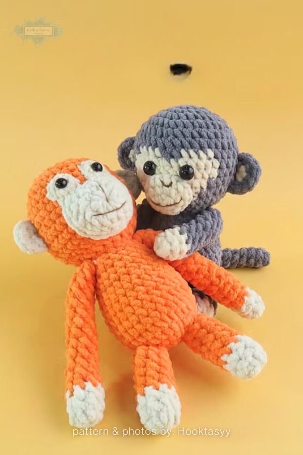 An image of two handmade crochet monkey plushies from a crochet roundup on craftinghappiness.com. The pair features one orange monkey lying down and one grey monkey sitting behind it, both made from chunky, textured yarn with cream-colored faces and paws. They have small black safety eyes and simple embroidered smiles, set against a solid yellow background.