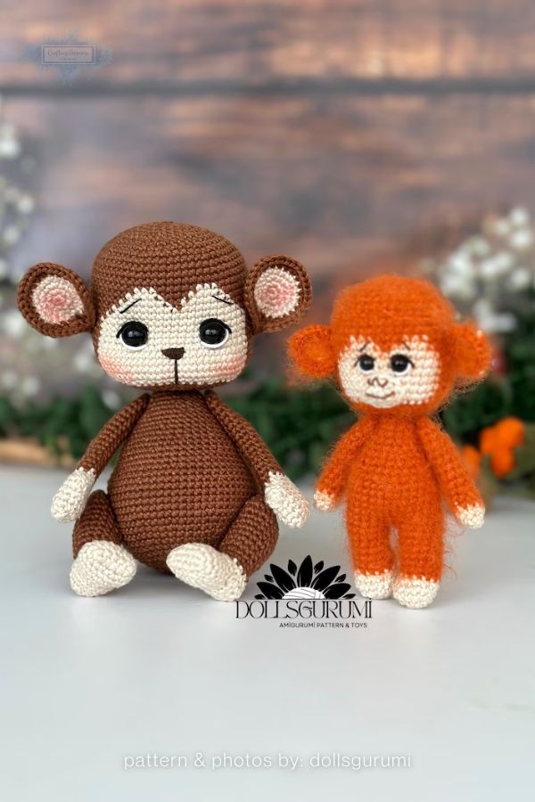 An image of two handmade crochet monkey plushies from a crochet roundup on craftinghappiness.com. The pair includes a larger brown monkey with a rounded body and pink-blushed cheeks, and a smaller, fuzzy orange monkey. Both have cream-colored faces and paws with detailed embroidered facial features and large, dark eyes, posed against a soft-focus background with wooden textures and greenery.