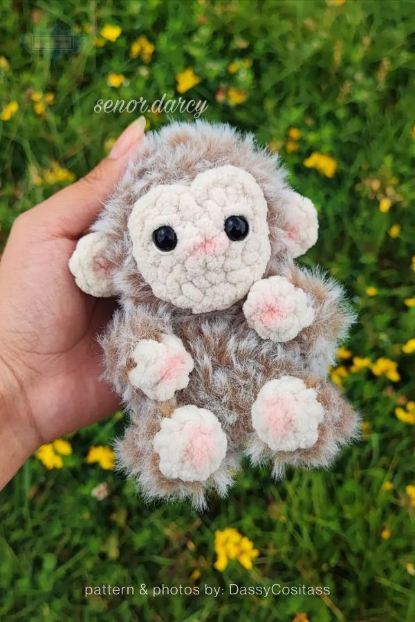 An image of a fuzzy, handmade crochet monkey plushie from a crochet roundup on craftinghappiness.com. A hand holds the monkey, which is made from textured, multi-tonal brown and grey faux-fur yarn. It features a cream-colored face, ears, and paws with soft pink accents on the nose and pads. The monkey has large black safety eyes and is set against a vibrant green outdoor background with yellow flowers.