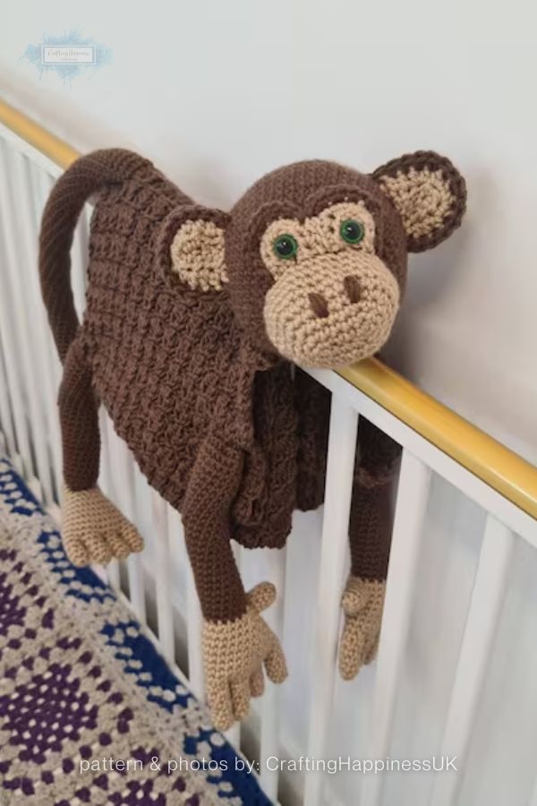 An image of a handmade crochet monkey rug from a crochet roundup on craftinghappiness.com. The monkey is crafted from dark brown yarn with tan accents on its face, inner ears, hands, and feet, and it features green safety eyes. It is shown draped over a white crib railing with its head resting on the top bar and its long limbs and tail hanging down.
