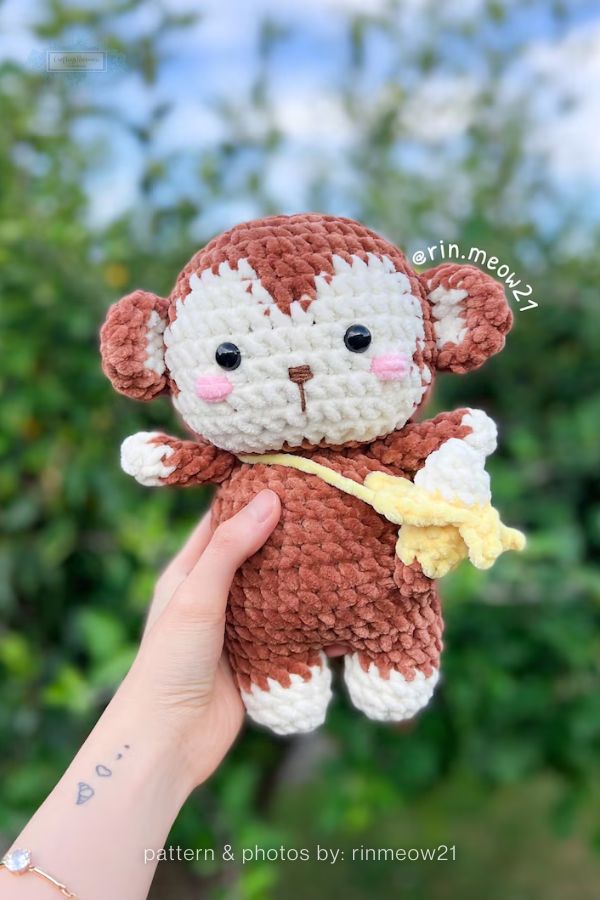 An image of a handmade crochet monkey plushie from a crochet roundup on craftinghappiness.com. A hand holds the monkey, which is made from plush brown and cream yarn and features a small yellow crochet bag slung across its body. The monkey has rosy pink cheeks, small black safety eyes, and an embroidered nose and mouth, set against a soft-focus outdoor background of green foliage and a blue sky.