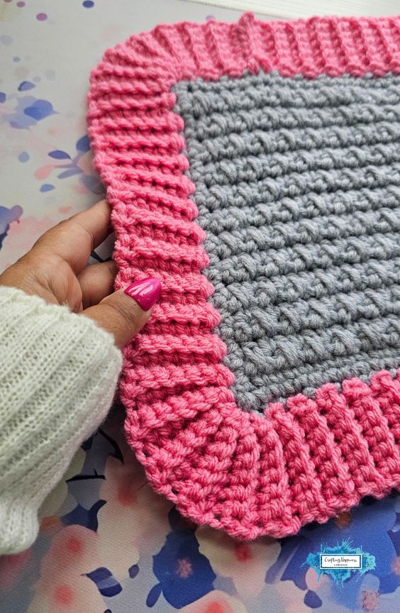 A hand with bright pink nails gently holding the corner of a completed grey crochet square that features a wide, textured pink ribbed border, set against a floral watercolor background. A small blue Crafting Happiness logo is in the bottom right corner. Original design by Ariana of CraftingHappiness.