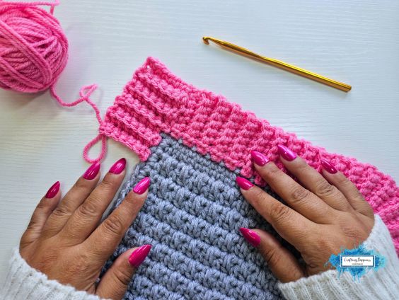 A top-down view of a person's hands with bright pink nails resting on a grey crocheted piece with a pink ribbed border. In the background, a ball of pink yarn sits on the left and a gold crochet hook sits on the right. A small blue Crafting Happiness logo is in the bottom right corner. Original design by Ariana of CraftingHappiness.