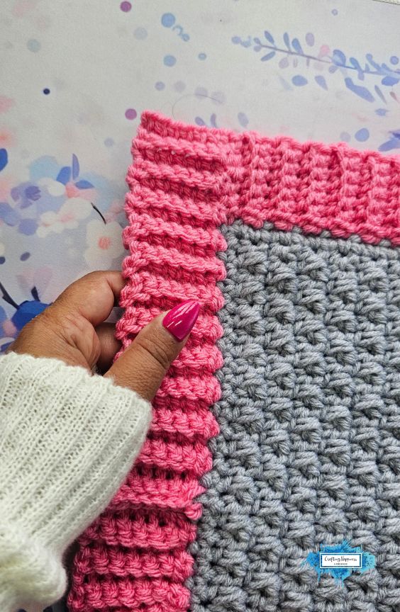 A vertical close-up shot showing a hand with pink nails lightly holding the edge of a pink ribbed crochet border on a grey textured square, resting on a pastel floral background. A small blue Crafting Happiness logo is in the bottom right corner. Original design by Ariana of CraftingHappiness.
