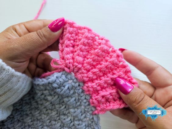 A close-up shot of a pair of hands with pink nails holding and demonstrating a section of the pink ribbed crochet border attached to a grey crochet project. A small blue Crafting Happiness logo is visible in the bottom right corner. Original design by Ariana of CraftingHappiness.