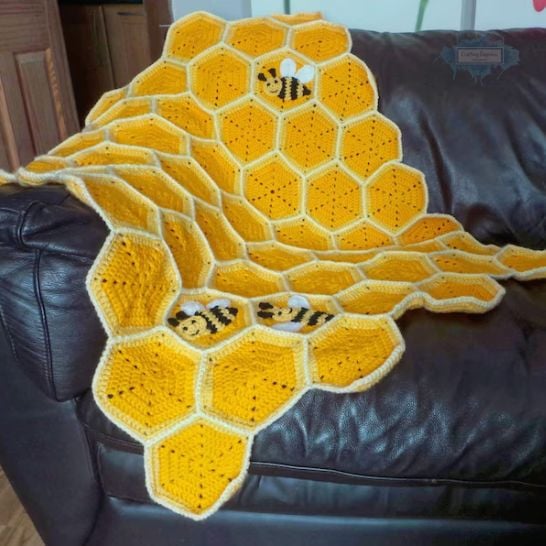 A bright yellow crochet blanket draped over a dark leather couch. The blanket is constructed from individual hexagonal motifs that form a honeycomb structure, complete with textured raised borders. Small, detailed crochet bumblebees with black and yellow stripes and white wings are appliquéd onto several of the hex tiles. This is a blanket pattern made by Ariana Wimsett of Crafting Happiness, a well known crocheter and author.