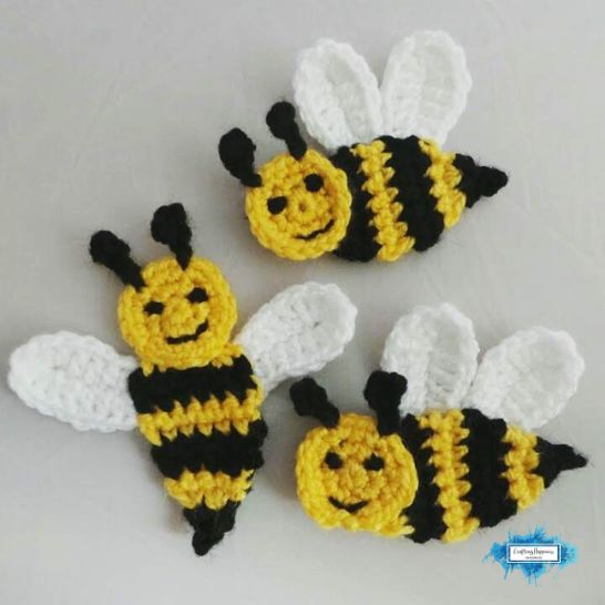 Three handmade crochet bumblebee appliques arranged on a plain light background. Each bee features a round yellow head with stitched black eyes and a smile, black antennae, a body with alternating yellow and black stripes, a pointed black tail, and two white textured wings.