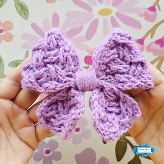 A person holding a textured crochet bow made from light purple yarn. The bow features an intricate, overlapping textured stitch pattern resembling a braided Celtic weave, with a smooth band of wrapped yarn neatening the center. The background shows a soft floral design.