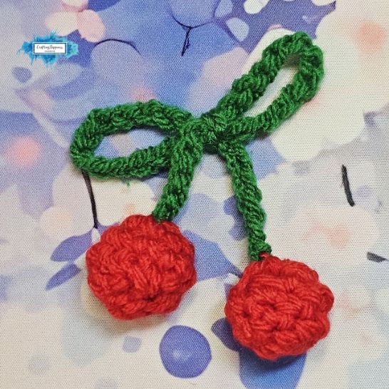 A crochet cherry embellishment lying flat against a blue and white patterned floral fabric. It consists of a long, thin green yarn loop forming a bow shape with two trailing stems, each ending in a bright red, plump textured crochet cherry.