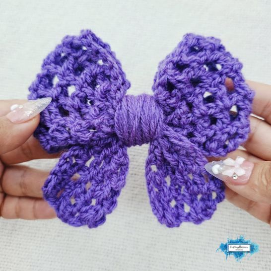 A purple crochet bow held by two hands to show its openwork structure. The bow is worked in a decorative mesh or lace-like stitch pattern with noticeable open spaces, finished with a thick section of matching purple yarn wrapped around the center.