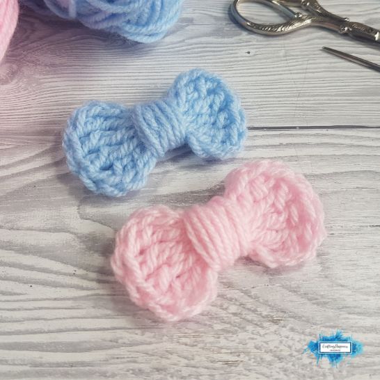 Two small, simple crochet bows placed side by side on a light-colored wooden surface. One bow is light blue and the other is light pink, both featuring a basic ribbed texture and a neat center wrapped tightly with matching yarn. Crafting tools are partially visible in the background.