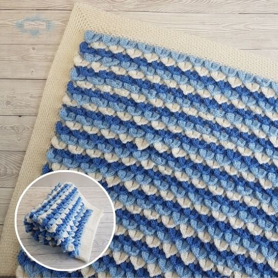 A detailed top-down view of a textured crochet blanket featuring layered, scale-like stitches that resemble rows of small overlapping leaves or hearts. The design alternates in horizontal stripes of white, light blue, and royal blue yarn with a clean, cream-colored ribbed border along the top edge. A circular inset shows the blanket completely folded on a light wooden floor. This is a blanket pattern made by Ariana Wimsett of Crafting Happiness, a well known crocheter and author.