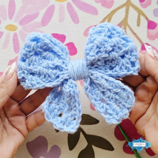A light blue crochet bow being held up against a colorful pink and purple floral background. The bow uses the classic cluster-based granny stitch pattern, creating small clusters of stitches with slight gaps, gathered in the middle with wrapped blue yarn.