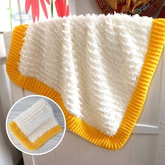 A textured white crochet baby blanket draped over a white wooden crib, featuring a thick, vibrant yellow ribbed border. The main body of the blanket consists of fuzzy, loopy horizontal ridges that give it a plush appearance. A circular inset in the lower left corner displays the blanket neatly folded to show off the contrast between the white textured center and the yellow frame. This is a blanket pattern made by Ariana Wimsett of Crafting Happiness, a well known crocheter and author.