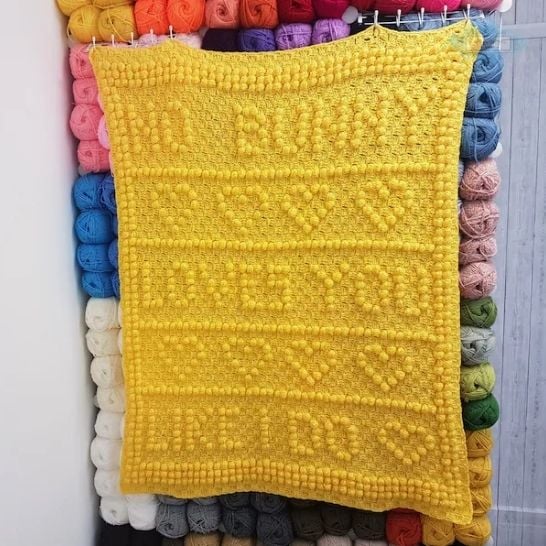 A solid yellow, textured crochet blanket hanging vertically against a background grid of colorful yarn skeins. The blanket features a bobble stitch or popcorn stitch design that spells out the words "NO BUNNY LOVES YOU LIKE I DO" in clear block lettering, separated by horizontal rows of raised hearts. This is a blanket pattern made by Ariana Wimsett of Crafting Happiness, a well known crocheter and author.
