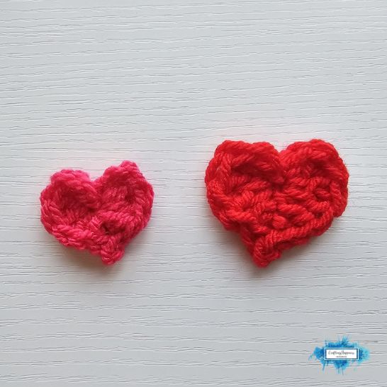 Two simple crochet heart appliques lying flat on a white wooden plank surface. On the left is a smaller bright pink heart, and on the right is a slightly larger vibrant red heart, both showing clean, defined stitches forming a classic heart shape.