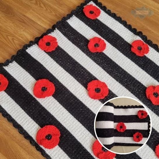 A striking crochet baby blanket featuring bold, alternating horizontal stripes of black and white yarn, spread flat over a wooden floor. The blanket is adorned with vibrant, three-dimensional red crochet poppy flowers with black centers scattered across the stripes, and it is finished with a delicate black scalloped border. A circular inset in the bottom right corner shows a section of the blanket rolled up, highlighting the contrast of the stripes and the layered floral details. This is a blanket pattern made by Ariana Wimsett of Crafting Happiness, a well known crocheter and author.
