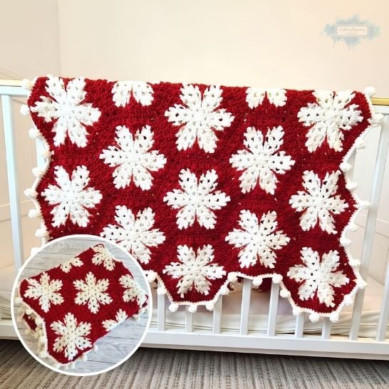 A winter-themed baby blanket draped over the side of a white wooden crib. The blanket features large, crisp white crochet snowflakes joined together over a rich, deep red background, finished with a white bobble or pom-pom border. A circular inset in the bottom left shows the red and white snowflake blanket neatly folded. This is a blanket pattern made by Ariana Wimsett of Crafting Happiness, a well known crocheter and author.