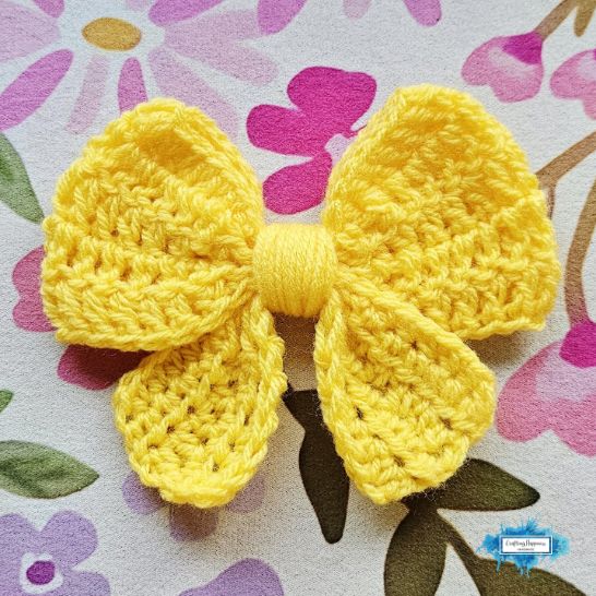A bright yellow crochet bow showcased against a pastel floral background. The bow is constructed using a solid, dense double crochet stitch pattern that gives it a full, uniform texture, complete with a neatly wrapped yarn center.