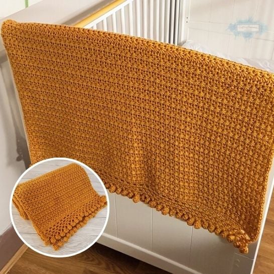 A textured, solid mustard-gold crochet blanket draped over the edge of a white crib. The stitch design consists of a dense, repeating star or flower-like cluster texture, finished with a row of small, matching crochet pom-poms along the bottom border. A circular inset in the lower left corner shows the blanket neatly folded, showcasing the uniform stitch work. This is a blanket pattern made by Ariana Wimsett of Crafting Happiness, a well known crocheter and author.