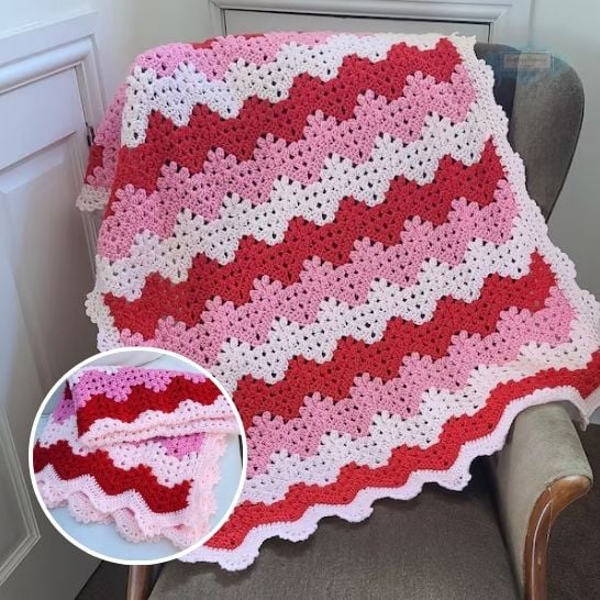 A vibrant chevron or ripple-stitch crochet blanket displayed over the back of an armchair. The design features alternating wavy stripes of deep red, bright pink, and clean white yarn, finished with a delicate pink scalloped edge. A circular inset in the lower left corner shows the blanket folded over to display its distinct zigzag texture. This is a blanket pattern made by Ariana Wimsett of Crafting Happiness, a well known crocheter and author.