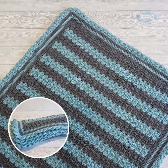 A close-up view of a textured crochet blanket with alternating horizontal stripes of charcoal gray and light aqua blue yarn, laid out flat on a light wood paneled floor. The blanket is completed with a matching aqua ruffled border. A circular inset in the lower left corner shows the blanket folded lengthwise to display the uniform texture of the stripes and the edge detailing. This is a blanket pattern made by Ariana Wimsett of Crafting Happiness, a well known crocheter and author.