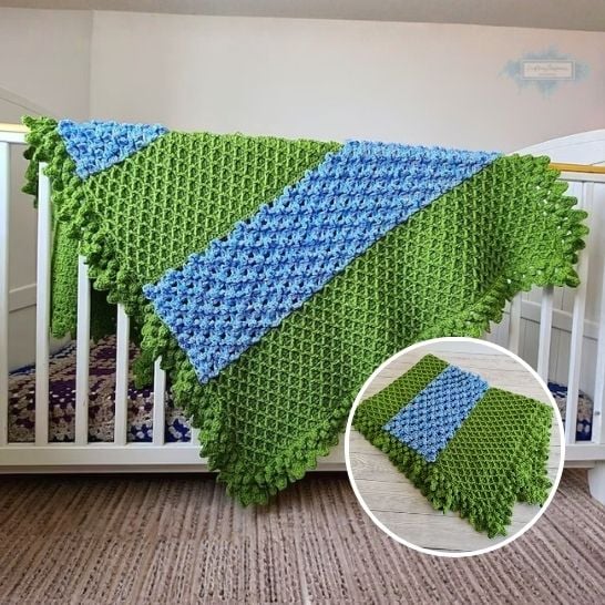 A green textured crochet blanket draped over a white wooden crib. The blanket features a prominent diagonal stripe of bright blue textured stitches running through the center, and the outer edges are finished with a thick, loopy green ruffle. A circular inset in the lower right corner displays the blanket laid out flat on a gray wood floor, showing the clean geometric lines of the blue stripe across the green trellis texture. This is a blanket pattern made by Ariana Wimsett of Crafting Happiness, a well known crocheter and author.