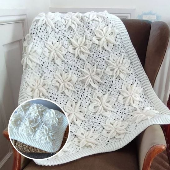 An elegant, monochromatic white crochet blanket draped over a brown leather armchair. The blanket is composed of joined square motifs, each featuring a large, raised 3D poinsettia flower with layered, pointed petals, and finished with a classic ribbed border. A circular inset in the lower left corner shows a close-up of the folded blanket resting on a textured surface, highlighting the intricate depth of the floral stitches. This is a blanket pattern made by Ariana Wimsett of Crafting Happiness, a well known crocheter and author.