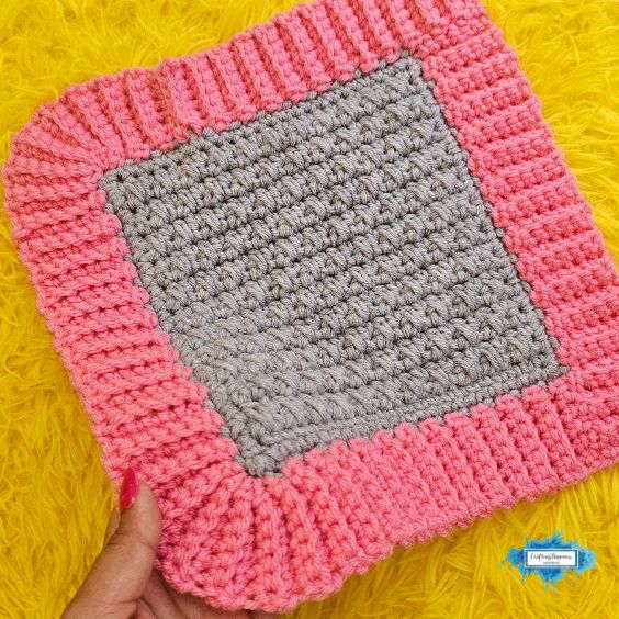 A completed square crochet project with a textured grey center and a pink ribbed border, lying flat on a bright yellow furry background. A small blue Crafting Happiness logo is in the bottom right corner. Original design by Ariana of CraftingHappiness.