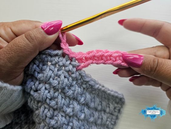 A close-up shot showing hands crocheting the very first row of the pink single crochet ribbing, working back down the foundation chain toward the grey project edge. A small blue Crafting Happiness logo is visible in the bottom right corner. Original design by Ariana of CraftingHappiness.