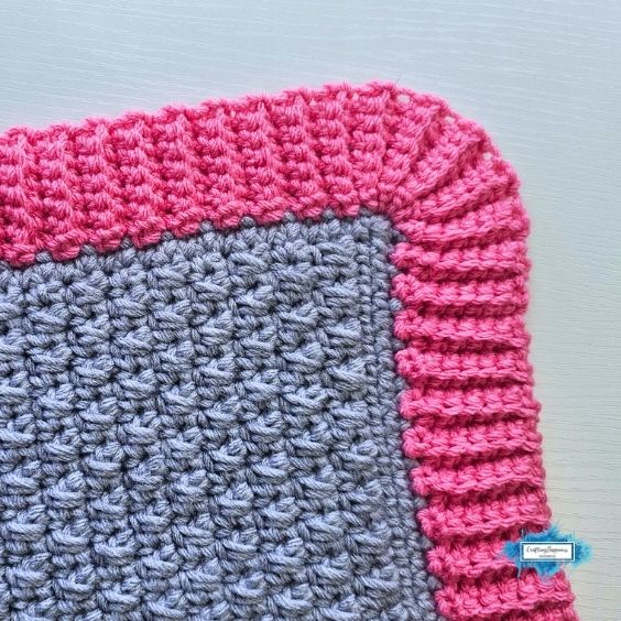 A close-up, top-down shot of a neatly crocheted project corner, showcasing a gently curved, rounded corner technique. The main body is textured grey, framed by a thick, vibrant pink single crochet ribbed border. A small blue Crafting Happiness logo sits in the bottom right corner. Original design by Ariana of CraftingHappiness.