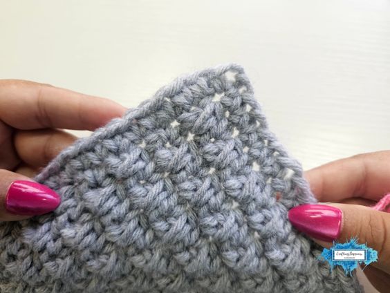 A close-up view of hands with bright pink manicured nails holding the edge of a completed single crochet grey project, demonstrating the base setup before adding a border. A small blue Crafting Happiness logo is in the bottom right corner. Original design by Ariana of CraftingHappiness.