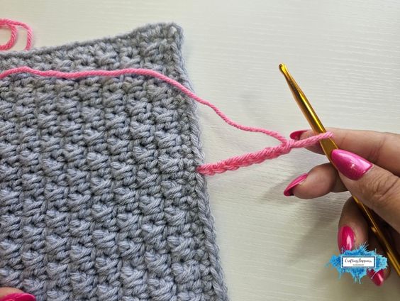 A hand holding a gold crochet hook attached to a short foundation chain of pink yarn that has been joined to the edge of a grey textured crochet piece. Original design by Ariana of CraftingHappiness.