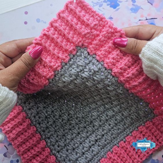 A close-up view of a person's hands with bright pink manicured nails gently stretching and holding open the corner of a crochet project to display a sharp, distinct 90-degree square corner. The center is textured grey surrounded by a pink ribbed border. A small blue Crafting Happiness logo is visible in the bottom right corner. Original design by Ariana of CraftingHappiness.