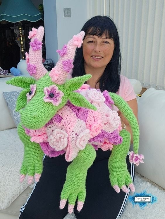 Ariana holding the completed 3-in-1 flower dragon crochet blanket in its plush dragon form, showing the green dragon body, flower-covered blanket, leafy details, and long dangling legs. Original design by Ariana of CraftingHappiness.