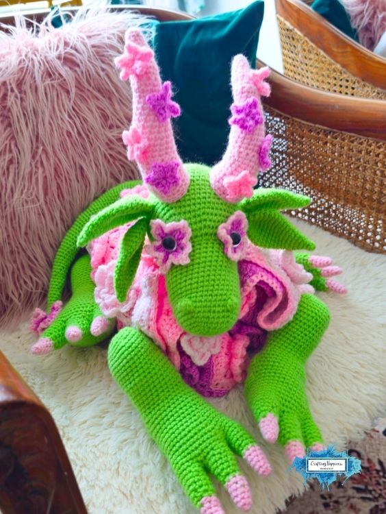Folded 3-in-1 flower dragon crochet blanket displayed as a plush dragon on a soft chair. The dragon features a green body, pink flower-covered blanket, leafy ears, floral horns, and long legs tucked around the folded blanket. Original design by Ariana of CraftingHappiness.