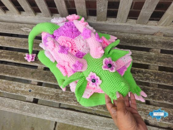 Top view of the folded flower dragon crochet blanket resting on a wooden bench, showing the dragon's face, floral horns, leafy ears, curled tail, and compact folded blanket shape. Original design by Ariana of CraftingHappiness.