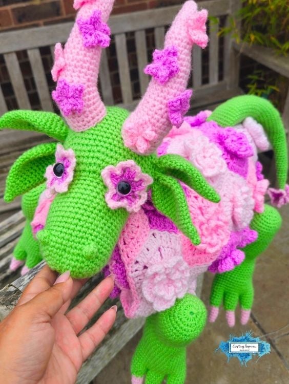 Close-up of the flower dragon crochet blanket draped over a garden bench, highlighting the dragon's face, flower-shaped eyes, leafy ears, floral blanket body, and textured crochet details. Original design by Ariana of CraftingHappiness.