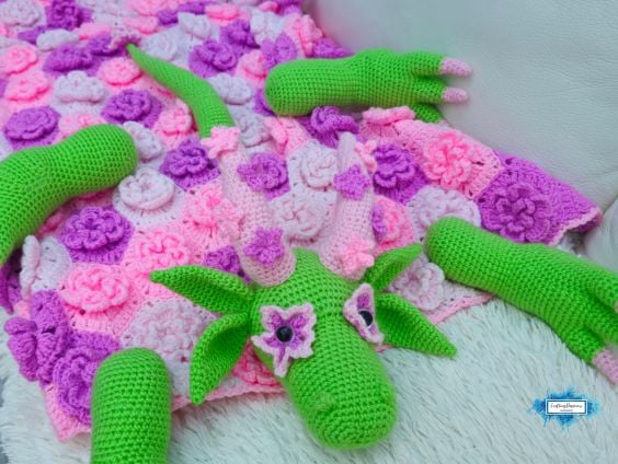The flower dragon crochet blanket opened flat to display the pink floral blanket with removable dragon head, legs, horns, and tail arranged around it. Original design by Ariana of CraftingHappiness.