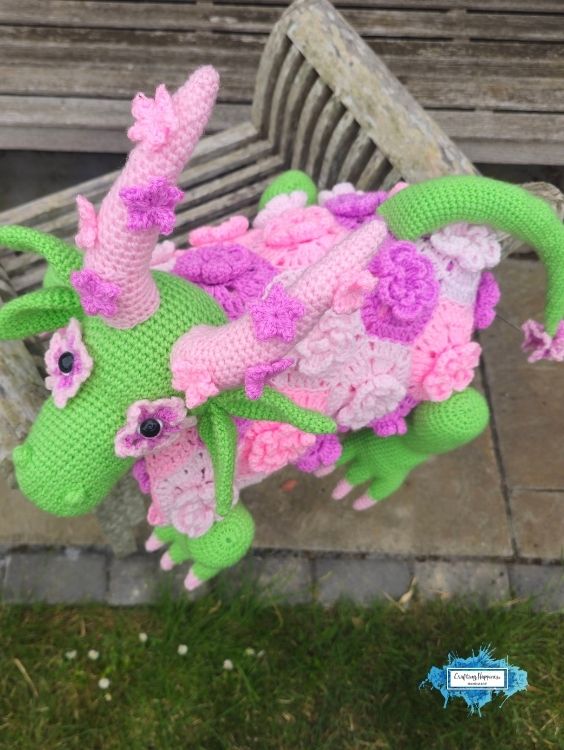Side view of the completed flower dragon crochet blanket displayed on a wooden bench, showing the raised floral blanket texture, long curled tail, leafy ears, and pink flower accents. Original design by Ariana of CraftingHappiness.