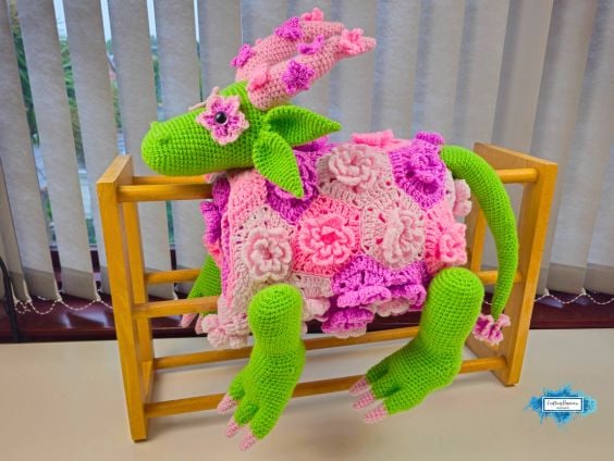 The completed flower dragon crochet blanket draped over a wooden blanket rack, featuring a green dragon body, layered pink crochet flowers, leafy ears, floral horns, and long legs hanging over the sides. Original design by Ariana of CraftingHappiness.