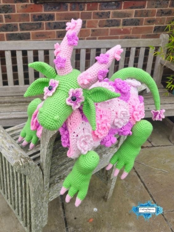 The finished flower dragon crochet blanket displayed across a wooden garden bench, featuring a green dragon with leafy ears, pink floral blanket body, curled tail, flower-shaped eyes, and long dangling legs. Original design by Ariana of CraftingHappiness.