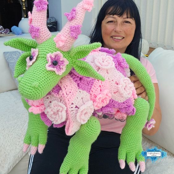 Ariana holding a large green and pink flower dragon crochet blanket in its plush dragon form, showing the leafy details, floral blanket, and long legs. Original design by Ariana of CraftingHappiness.