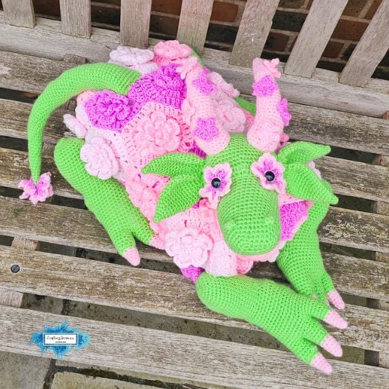 Folded flower dragon crochet blanket resting on a wooden bench, showing the green dragon head, leafy ears, floral blanket body, curled tail, and long front legs. Original design by Ariana of CraftingHappiness.