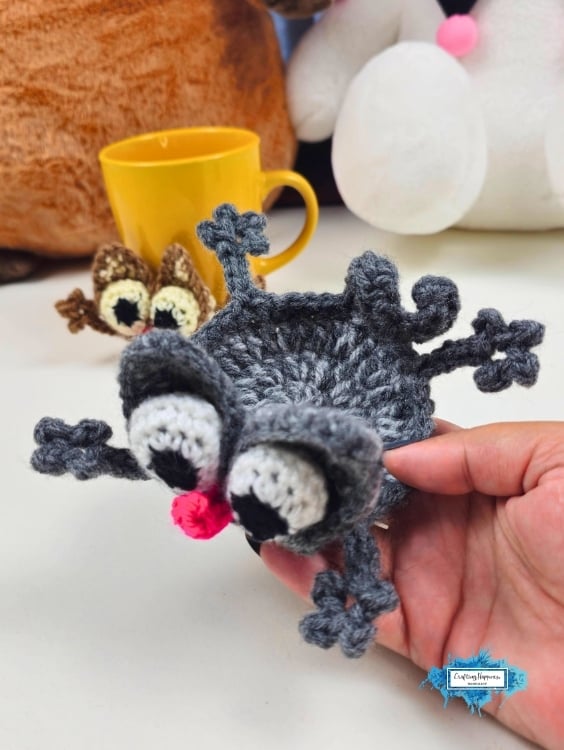 Close-up of a gray crochet splattered cat coaster held in one hand, showing its round body, oversized eyes, floppy ears, pink nose, and spread-out paws. A brown version and a yellow mug are softly blurred behind it. Original design by Ariana of CraftingHappiness.