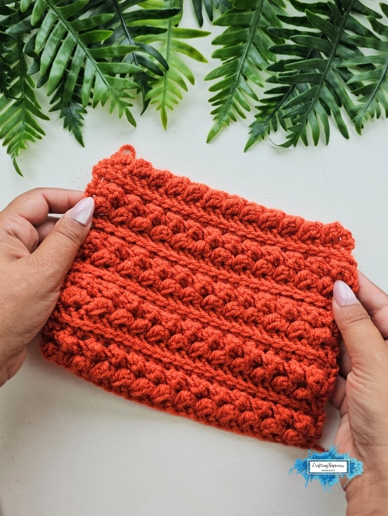 An orange crochet swatch featuring repeating raised rows of the Braided Leaves Stitch is held in both hands beneath green leafy foliage. The Braided Leaves Stitch was designed by Ariana Wimsett, a known crocheter and author, and the crochet work shown here was made by her.