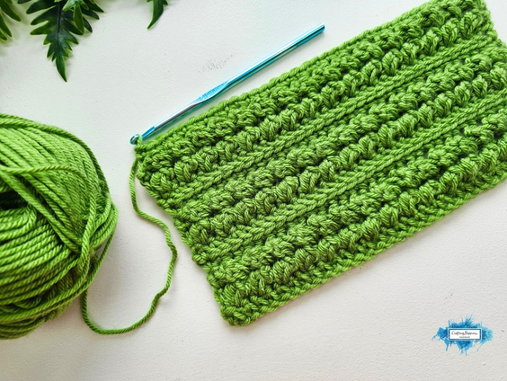A green crochet swatch featuring repeating textured rows of the Braided Leaves Stitch sits beside a matching yarn ball and blue crochet hook. The Braided Leaves Stitch was designed by Ariana Wimsett, a known crocheter and author, and the crochet work shown here was made by her.